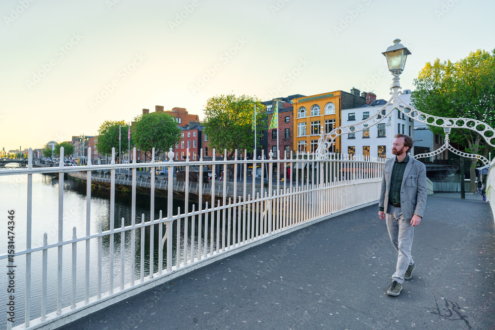 Naklejka premium A man walks across Dublin’s iconic Ha’penny Bridge at sunset, with colorful riverside buildings and calm waters in the background.