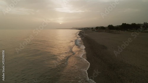 Drone image of a warm glowing sunset over the beach and sea near Rethimno, Crete, Greece showing reflections in the water and empty sunbeds along the shore