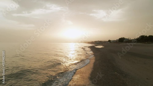 Aerial view of a serene empty beach near Rethimno, Crete, Greece with the sun setting over calm sea waters and soft waves reaching the shore under a cloudy sky