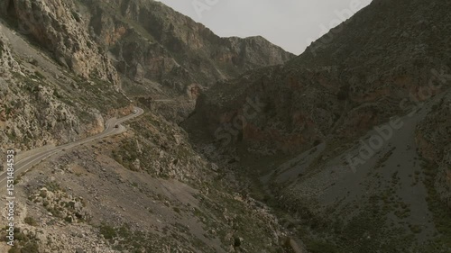 Aerial view of a narrow paved road snaking along steep cliffs and rocky landscape in Kourtaliotiko Gorge, Crete, Greece, under a hazy sky with minimal vegetation
