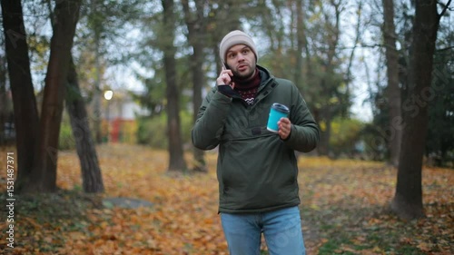 A man in a green jacket and beige beanie walks through a park in autumn, talking on the phone and holding a blue coffee cup. He appears relaxed in a casual outdoor setting.
