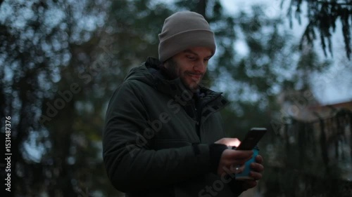A young man in a green jacket and beige beanie stands in a park on a chilly day, smiling while looking at his phone and holding a blue coffee cup. Autumn atmosphere and relaxed mood.
