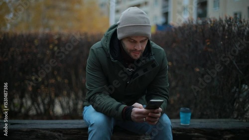 A man in a green jacket and beige beanie sits on a park bench in autumn, focused on his smartphone. A reusable coffee cup rests beside him as he scrolls or types on his phone.