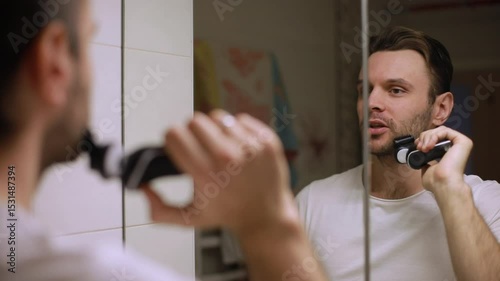 A young man grooming his beard with an electric trimmer in a modern bathroom. He looks into the mirror with focus, wearing a plain white t-shirt. Morning hygiene routine concept.
