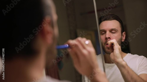 A young man in a white t-shirt brushes his teeth with an toothbrush while looking in the bathroom mirror, starting his morning hygiene routine in a home setting.
