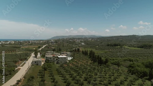 Drone view over olive and lemon tree orchards near Asprouliani, Crete, showing a scenic mix of farmland, village houses, winding road and coastal background under blue sky