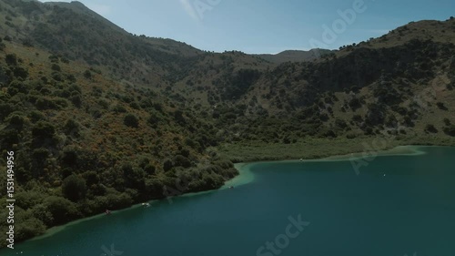 Top view of a wooden dock nestled in trees beside the vivid shallows of Lake Kournas, where a small white boat floats peacefully in Crete, Greece