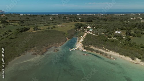 Narrow inlet connects to Lake Kournas where colorful pedal boats are docked along the beach with surrounding vegetation, small pier, and distant countryside