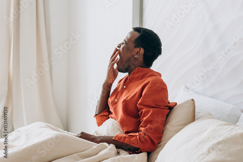Black man sitting in bed, yawning while covering his mouth with his hand, waking up in the morning and feeling the weight of tiredness and sleepiness lingering