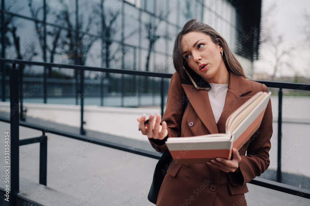 Fototapeta premium Businesswoman talking on phone and reading book outdoors