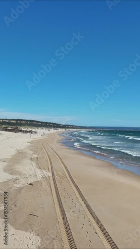 High drone view of Caparica beach near Lisbon, Portugal. Vehicle tracks drawn in the sand meet soft Atlantic waves. Aerial shot showing natural patterns and contrast between land and sea.

