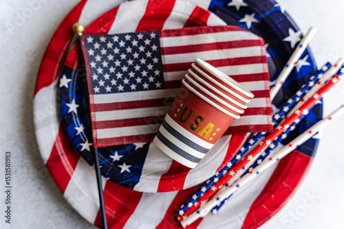 Overhead view of a stack of paper cups, paper plates, drinking straws and an american flag on a table for Independence Day on fourth of July photo