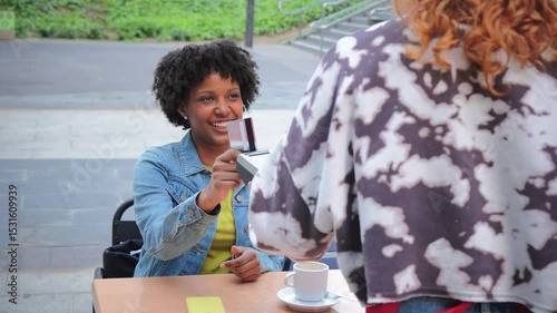 A cheerful woman is engaging in a payment transaction with a credit card while seated at a table in a bustling cafe, showcasing the convenience of modern payment methods in social settings.