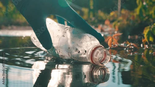 Hand in blue latex glove collecting plastic bottle from water puddle with sun backlight and lens flares slow motion closeup
