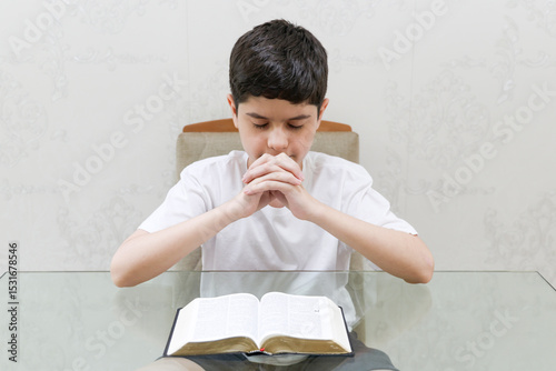 Brazilian preteen boy prays quietly with his hands clasped and head bowed in front of an open Bible. Eyes closed, he wears a white shirt and sits at a table indoors