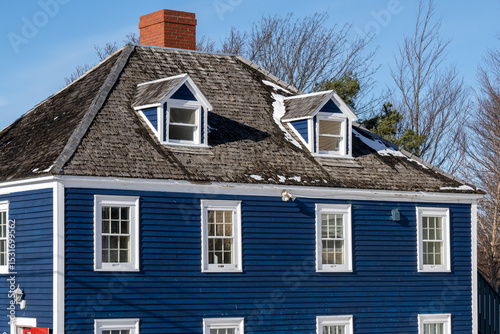 Vintage Mansard style roof with two dormer windows, cedar shake roof, red brick chimney, navy blue horizontal clapboard, double hung windows, and white trim siding on the antique building.