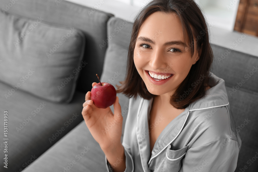 Beautiful young woman with fresh apple sitting on sofa in living room Beautiful young woman with fresh apple sitting on sofa in living room