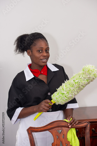 Domestic worker in black-and-white uniform holding a green duster and cleaning a wooden chair in a tidy indoor setting.