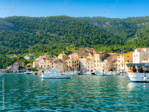 Charming harbor view of Komiža, Croatia, with colorful boats, stone houses, and green hills under a clear blue sky. 