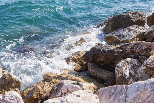 Fototapeta Naklejka Na Ścianę i Meble -  Rocky shore with turquoise sea water rolling in waves and creating splashes and thick white sea foam.