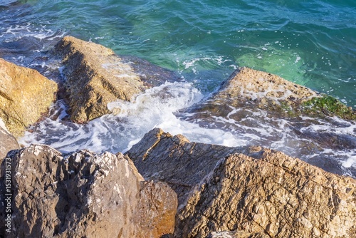 Fototapeta Naklejka Na Ścianę i Meble -  Rocky shore with turquoise sea water rolling in waves and creating splashes and thick white sea foam.