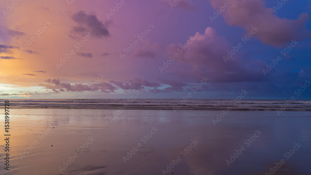 Obraz premium Sunset over the ocean. Pastel shades. Purple and pink clouds in a blue sky. Orange illumination near the horizon. The ocean waves roll towards the shore, spreading on the beach. Reflection on wet sand