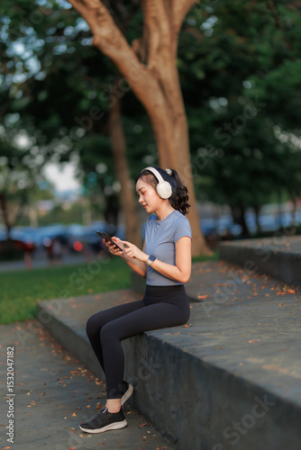 A young woman takes a break after exercising, relaxing with a cheerful smile. She wears headphones, listens to music, and chats with her friend on the phone, enjoying the peaceful moment.