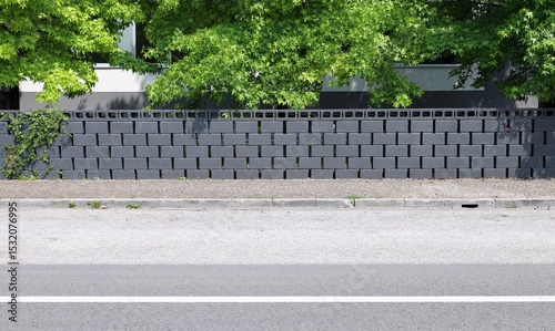 Fototapeta Naklejka Na Ścianę i Meble -  Dark gray concrete block fence, with every single piece separated from each other by a small space. Trees and modern building on behind. Sidewalk and street in front. Background for copy space.