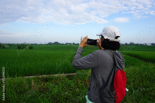 Woman using phone camera taking picture of the green paddy field with rural view background. Smartphone photographer taking nature landscape photos.