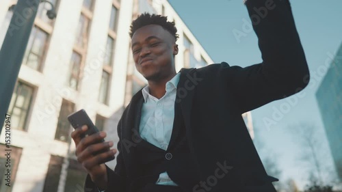 Portrait of joyful black guy looking at smartphone screen, rejoicing good news. Smiling african american person in city center, passerby in downtown, successful business deal, positive emotions
