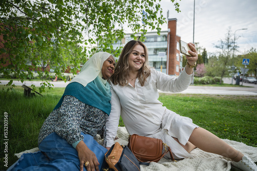 Two ethnically diverse women taking a selfie on picnic blanket