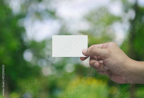 Man hand holding blank white business name card in fingers over blur green tree in park