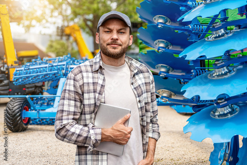 Male farmer with tablet standing next to farm implements at agricultural equipment dealership