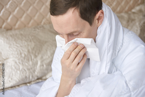 Caucasian adult male with cold or allergies blowing nose into tissue in bedroom wearing white robe.