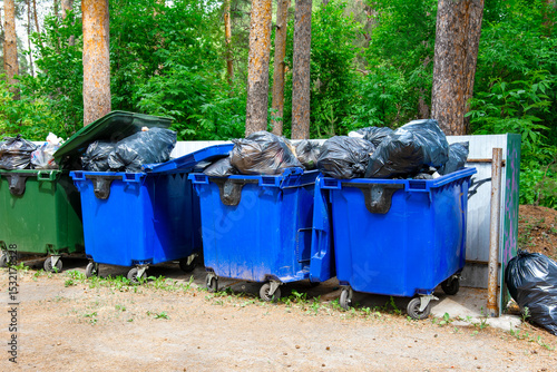 A row of overflowing dumpsters stands by the fence in a public park, causing environmental pollution.