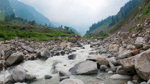 4K shot of Beas river flowing in Himalaya mountains in Solang Valley, Himachal Pradesh, India. Flowing river stream and campsite in distant at Palchani Thatch. Travel, trekking and vacation concept