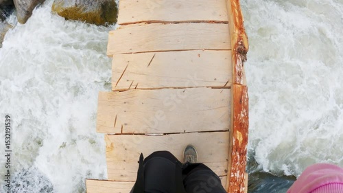 4K POV of young woman walks on a wooden bridge over a mountain river. Top view of woman legs in casual hiking boots. Adventure in nature. Tourist crossing small wooden bridge over purest Beas river