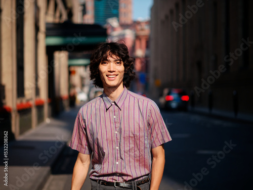 Portrait of handsome Chinese young man wearing stripe shirt posing in the street, young guy with black curly hair with urban background. Male fashion, cool Asian young man lifestyle.