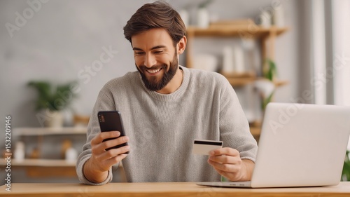 Young man holding a credit card and smartphone, completing an online transaction while sitting at a desk with a laptop in a cozy home.