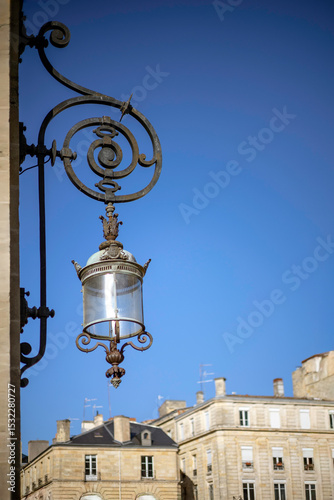 Ornate metal lantern with curved decorative ironwork and a cylindrical glass enclosure on the facade of Bordeaux City Hall (Hotel de ville de Bordeaux). Bordeaux, Gironde, Nouvelle-Aquitaine, France.
