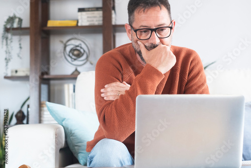 Mature man working from home on laptop, wearing orange sweater. Middle-aged freelancer using technology in domestic workspace. Various expressions from happy to thoughtful. Remote work, focus and life