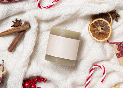 Green pillar candle with blank label on white knitted sweater near Christmas decor, winter mockup