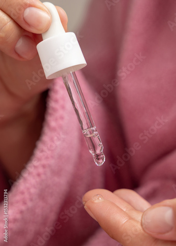 Young woman in pink bathrobe dropping essential oil on a finger and holding a pipette closeup