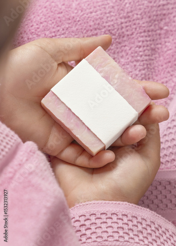 Closeup of young woman in pink bathrobe holding soap bar with blank label, mockup