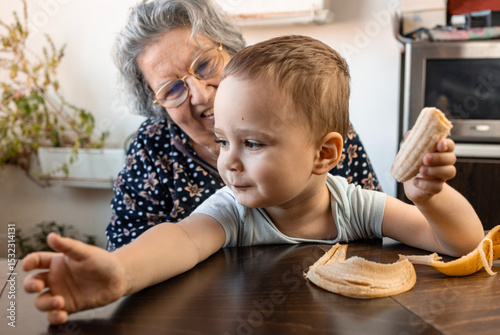 2,5 YO Jewish child holding banana sitting in his 80 year old grandmother on the table, smiling..