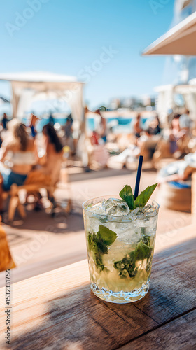 Glass of mojito cocktail with mint leaves and ice cubes on wooden bar counter in a beach club, with blurred people in the background enjoying summer vacation