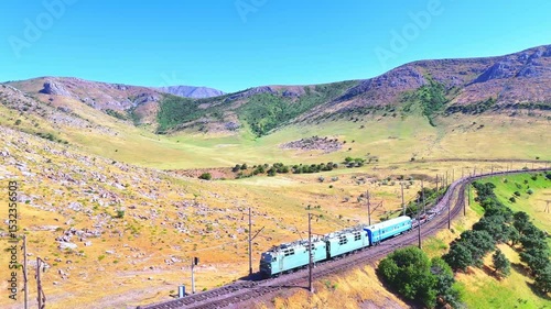 Freight train winding through mountain railway in Southern Kazakhstan. Rocky hills and green-yellow steppe form a scenic landscape.