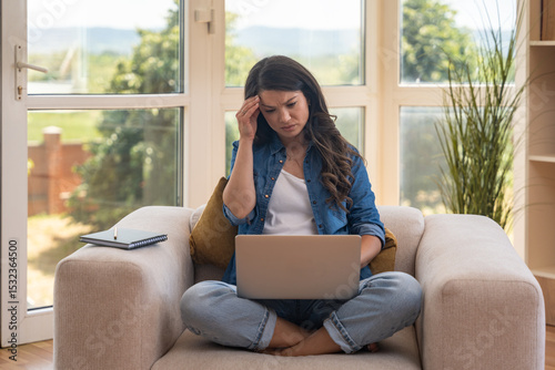 Young businesswoman working remotely from home, sitting on a cozy armchair with a laptop. Modern workspace, digital job, comfortable environment, and flexible online career opportunity
