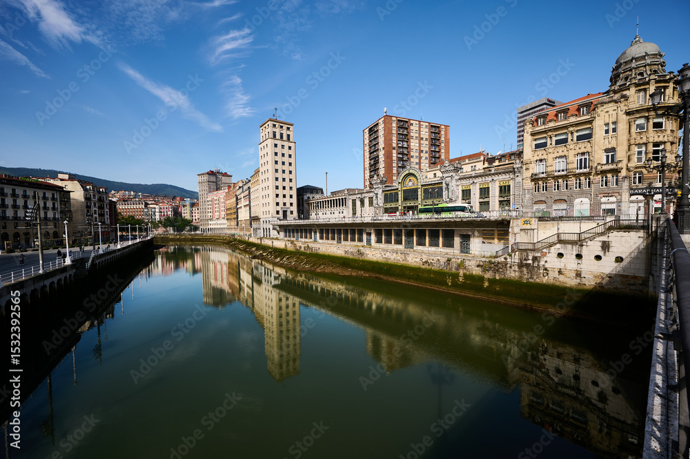 Obraz premium Cityscape of Bilbao shimmering in the Nervion river, featuring stunning reflections under a bright blue sky filled with clouds