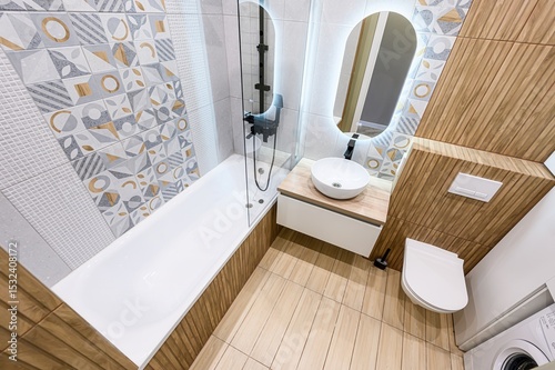 modern bathroom with a tub, vanity, toilet, and washing machine. It features patterned tiles, wooden accents, and a glowing oval mirror